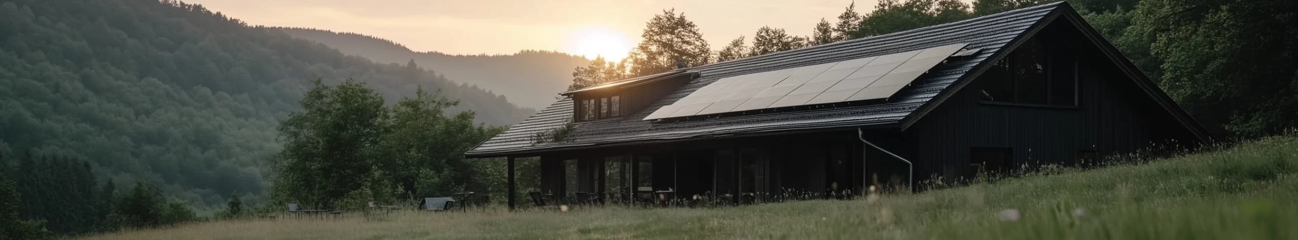 Hus med solpaneler på taket, omgivet av träd och kullar, vid solnedgången.