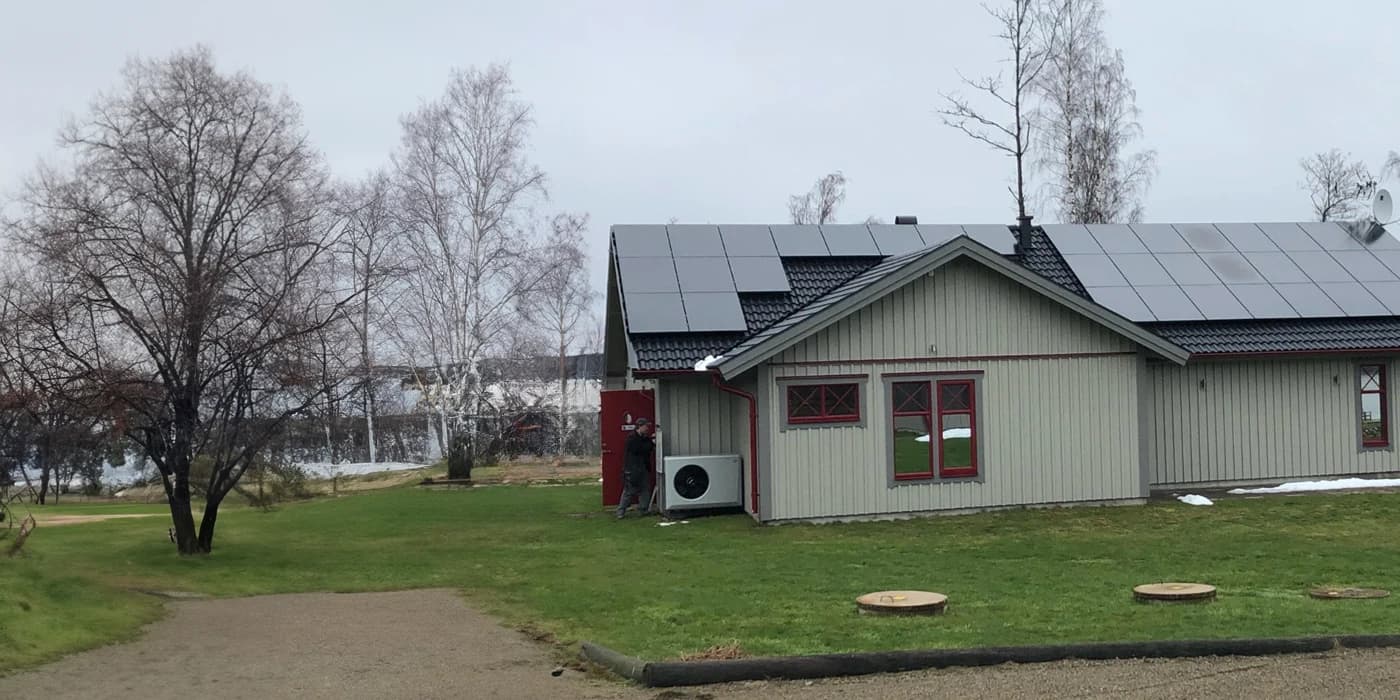 Ett litet grått hus med solpaneler på taket och rödkantade fönster, omgivet av gräs och lövlösa träd en molnig dag.