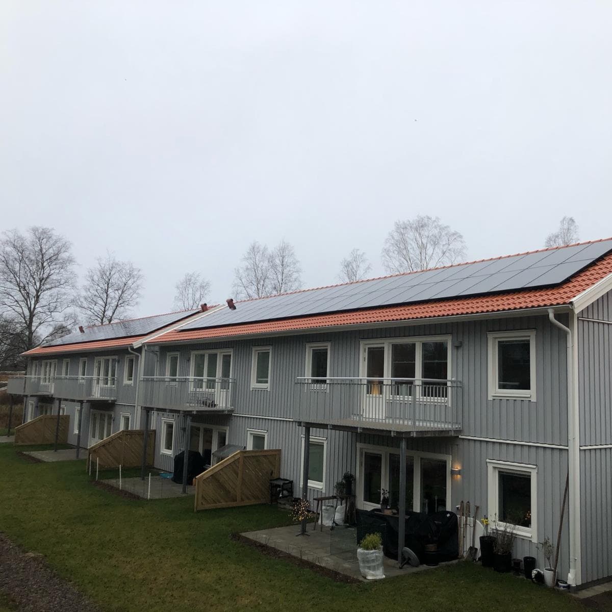 En rad moderna tvåvåningshus med grå fasad och orange tak, utrustade med solpaneler. Omgivningarna inkluderar gräs och kala träd under en mulen himmel.
