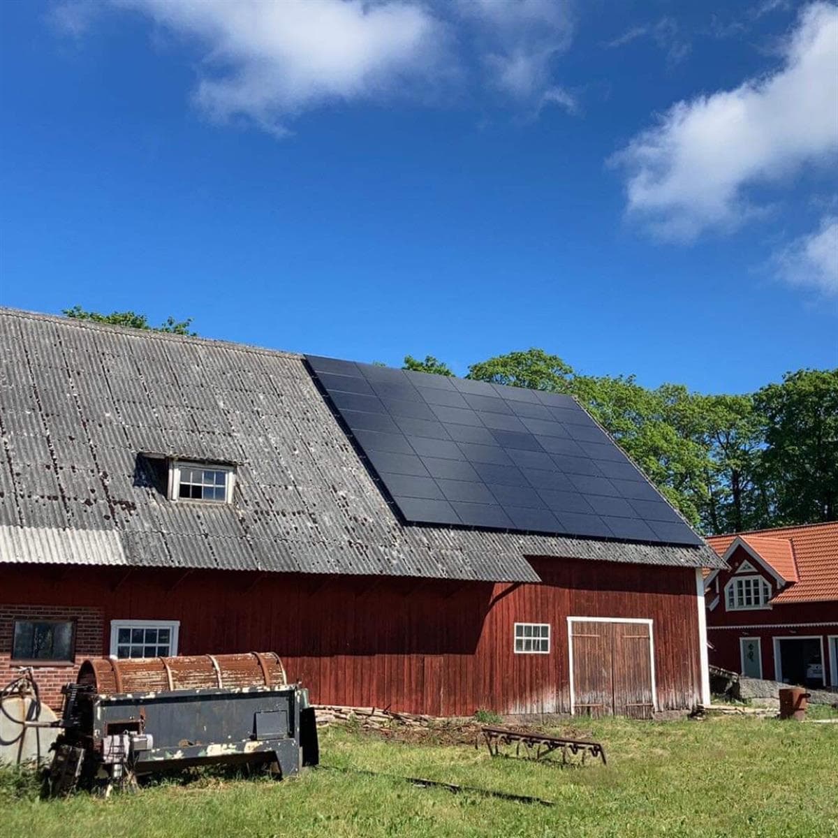 Solpaneler på ett sluttande tak på en röd lada med en blå himmel och några moln i bakgrunden. En vintage jordbruksmaskin är i förgrunden.
