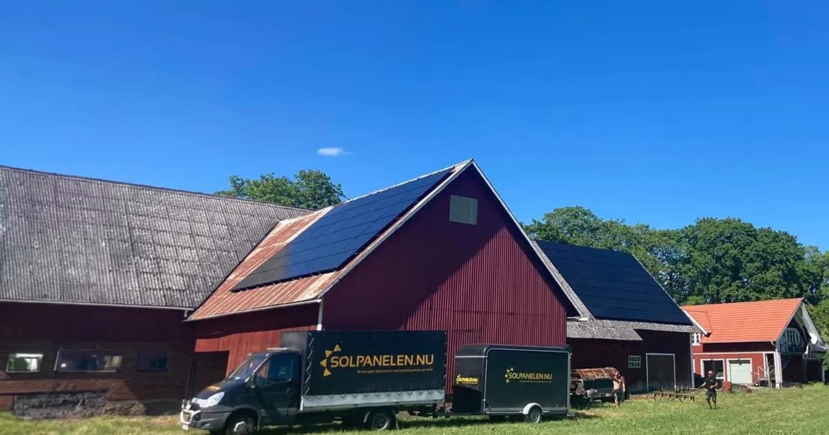 En röd ladugård med solpaneler på taket ligger inbäddad bland gröna träd, medan en lastbil och släpvagn med Solpanelen.nu sitter framför. Under en klarblå himmel återspeglar denna scen fördelarna med Solcellsbidrag för ett hållbart boende.