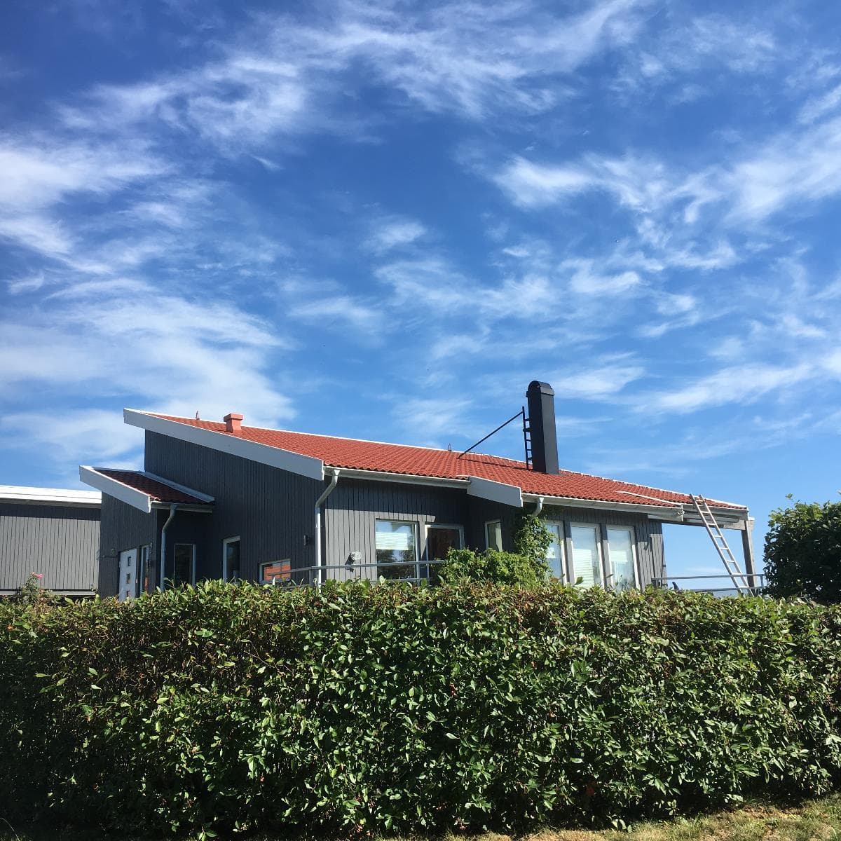 Grå villa med rött tak och solcellsanläggningen delvis synlig bakom häcken
