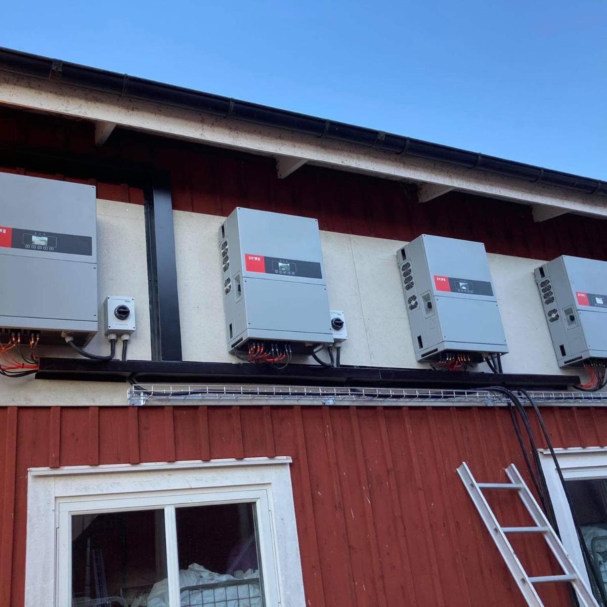 Tre elektriska växelriktare monterade på en röd vägg ovanför ett fönster, med kablar löpande och en stege lutad mot väggen.