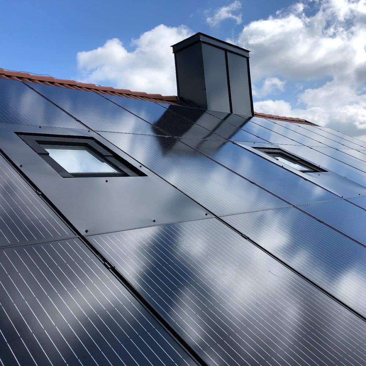 Solpaneler installerade på ett sluttande tak med en skorsten, reflekterande moln under en delvis molnig himmel.