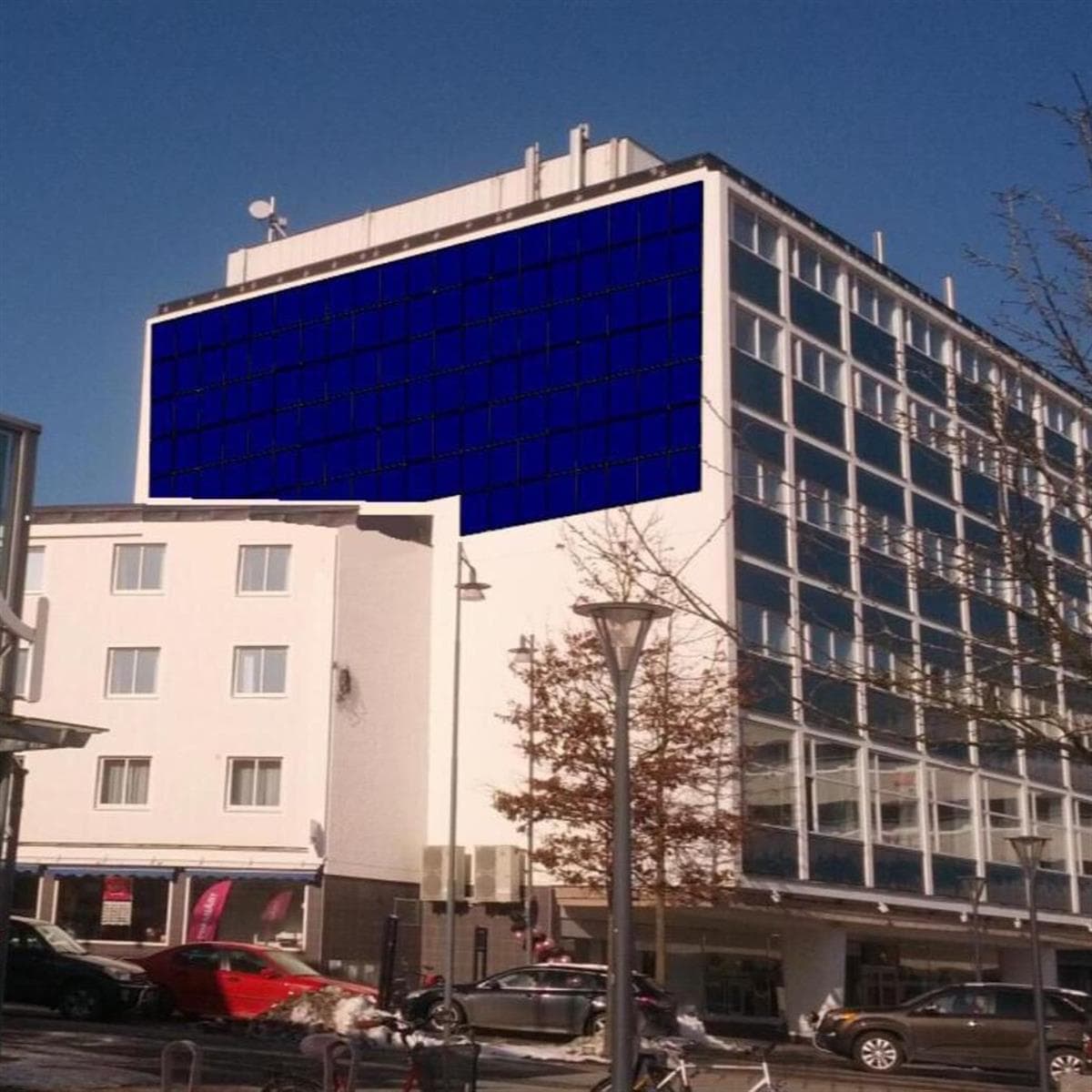 En modern vit byggnad med mörkblå paneler på de översta våningarna, i anslutning till en mindre vit byggnad, under en klarblå himmel.