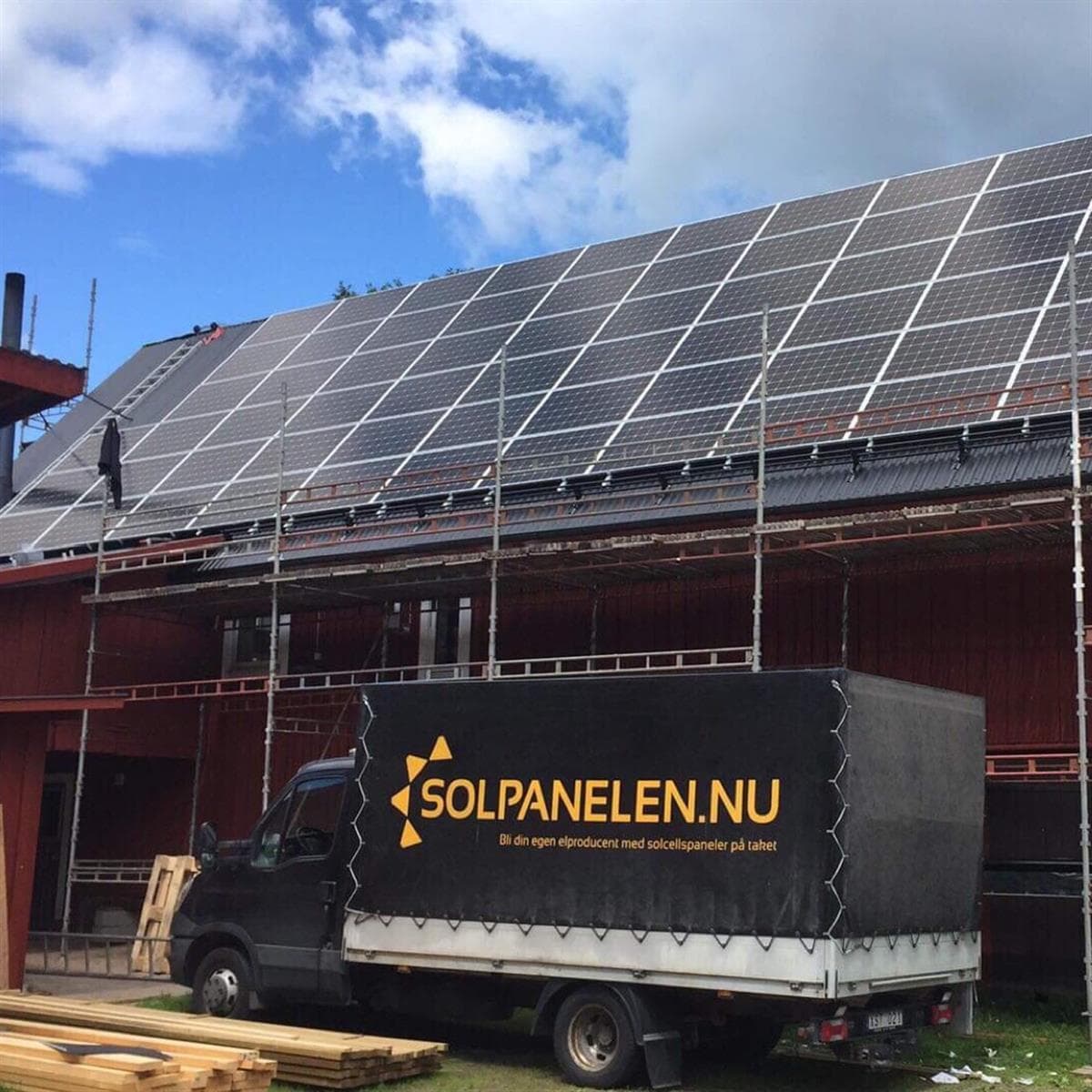 En lastbil märkt Solpanelen.nu står parkerad framför en byggnad med solpaneler som monteras på taket under delvis molnig himmel. Byggnadsställningar sätts upp runt byggnaden.