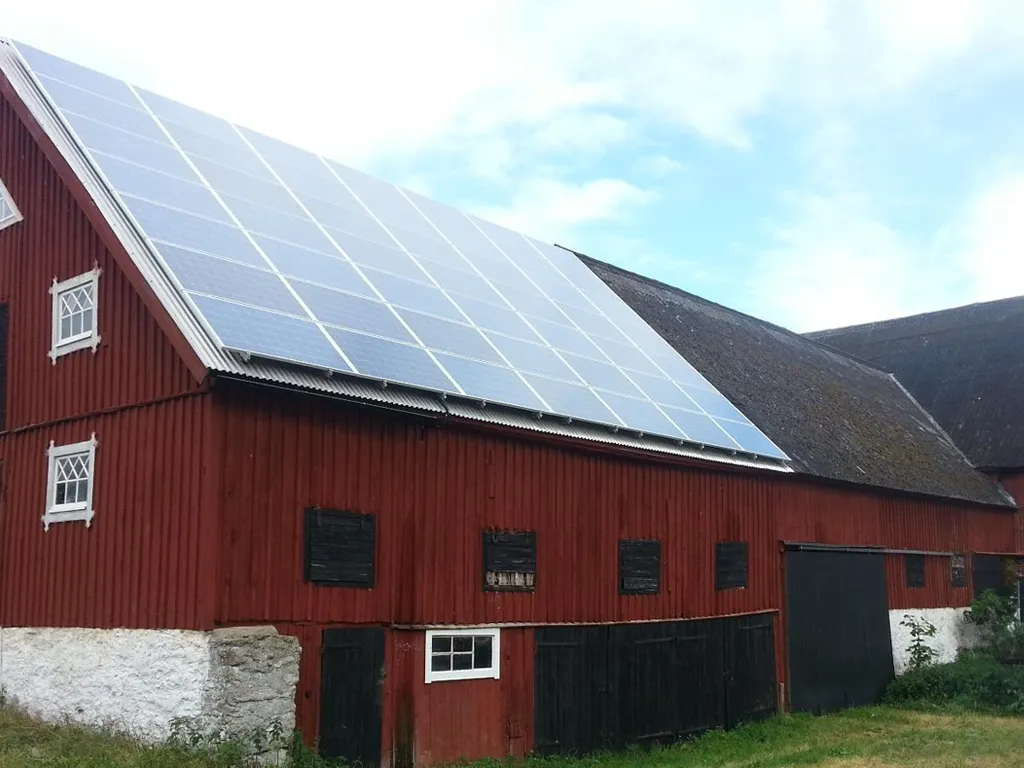 Röd lada med stora solpaneler installerade på taket, under en delvis molnig himmel.