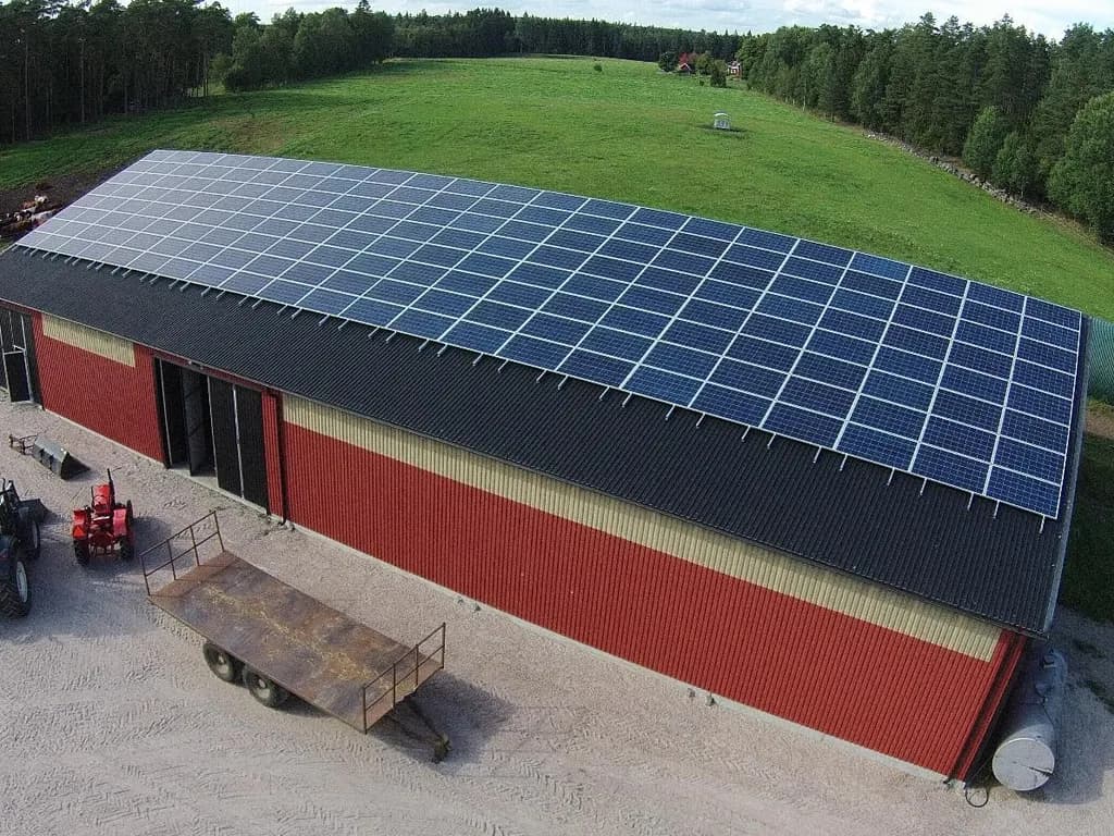 En stor, rektangulär röd ladugård med svart tak är utrustad med många solpaneler. Det ligger på landsbygden, omgivet av gräs och träd. En traktor och släp är parkerade i närheten.