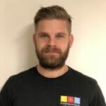 En skäggig man med kort hår iklädd en svart t-shirt står mot en vanlig bakgrund.