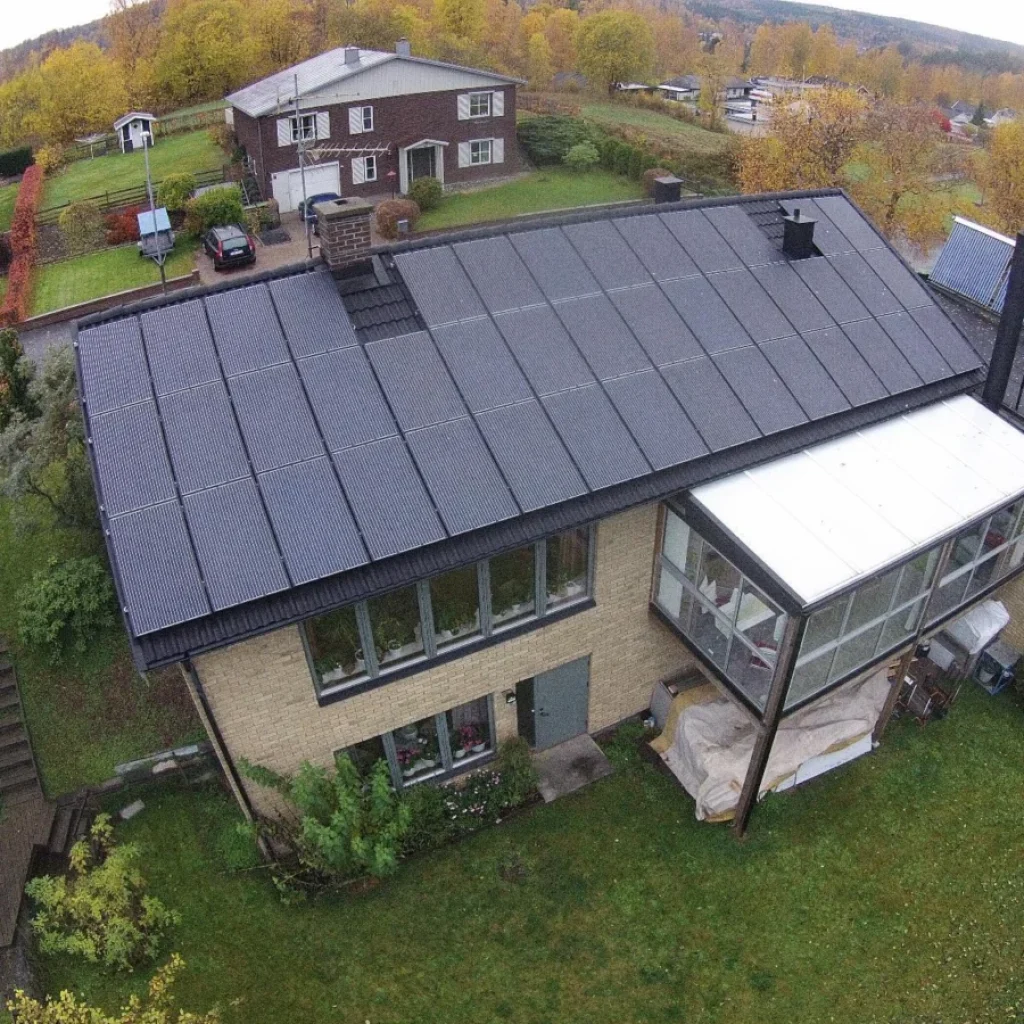 Flygfoto över ett hus med en stor solcellspanel på taket, en inglasad veranda och omgivande grönska en mulen dag.