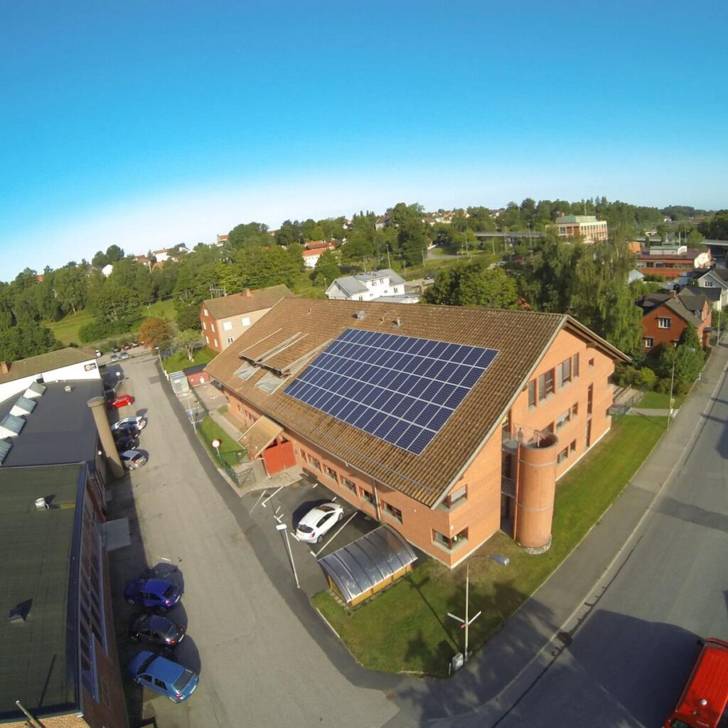 Flygfoto över en tegelbyggnad med solpaneler på taket, omgiven av träd, vägar och parkerade bilar under en klarblå himmel.