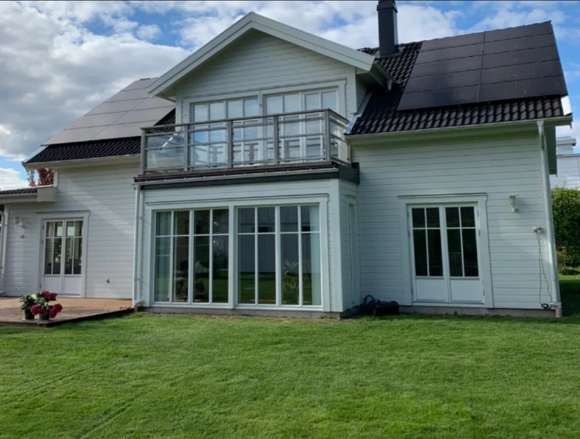 Vitt tvåvåningshus med glasbalkong, stora fönster, solpaneler och en grön gräsmatta under en halvmolnig himmel.