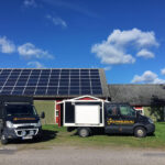 Två svarta lastbilar parkerade framför en byggnad med solpaneler på taket, under en blå himmel med moln.