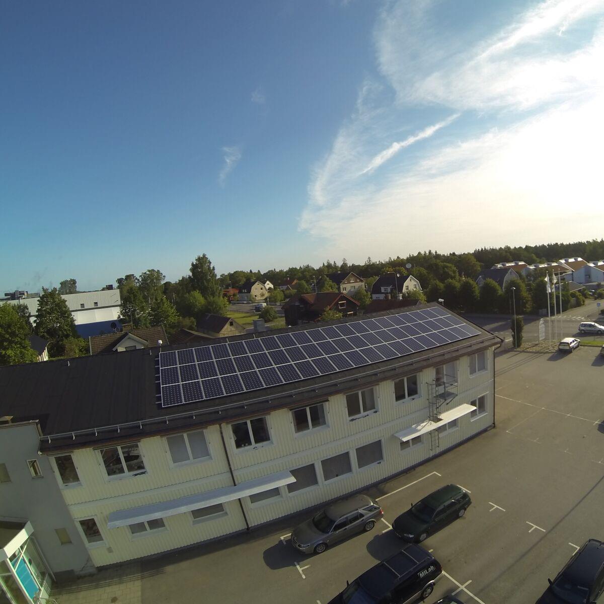 Flygfoto över en byggnad med solpaneler på taket. Flera bilar står parkerade på intilliggande tomt. Bostadsområde och träd syns i bakgrunden under en klar himmel.