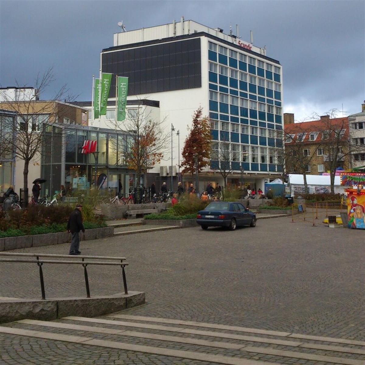 Stadtorg med stenbeläggning, en parkerad bil, människor som går och en modern byggnad med flera butiker, inklusive H&M. Mulen himmel i bakgrunden.