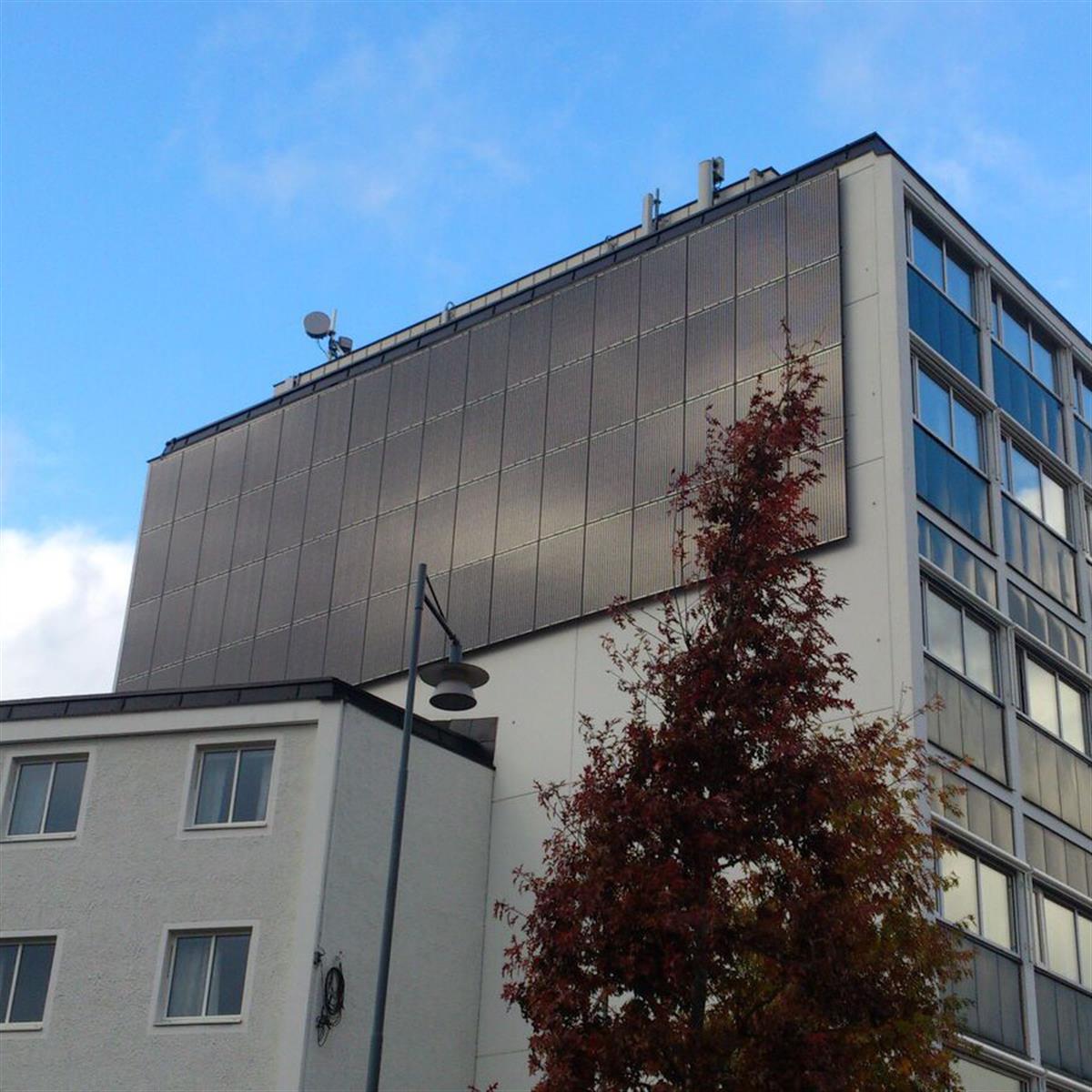 Byggnadens exteriör med en stor vertikal solpanelsuppsättning på den övre fasaden, i anslutning till mindre fönster. En gatlykta och ett träd med höstlöv är i förgrunden.