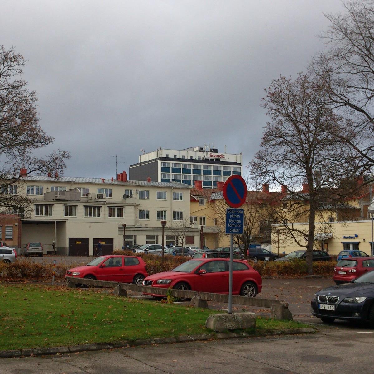 Mulet himmel ovanför en stadsbild med parkerade bilar, en parkeringsförbudsskylt och en stor byggnad med Scandics logotyp i fjärran. Träd utan löv är synliga.