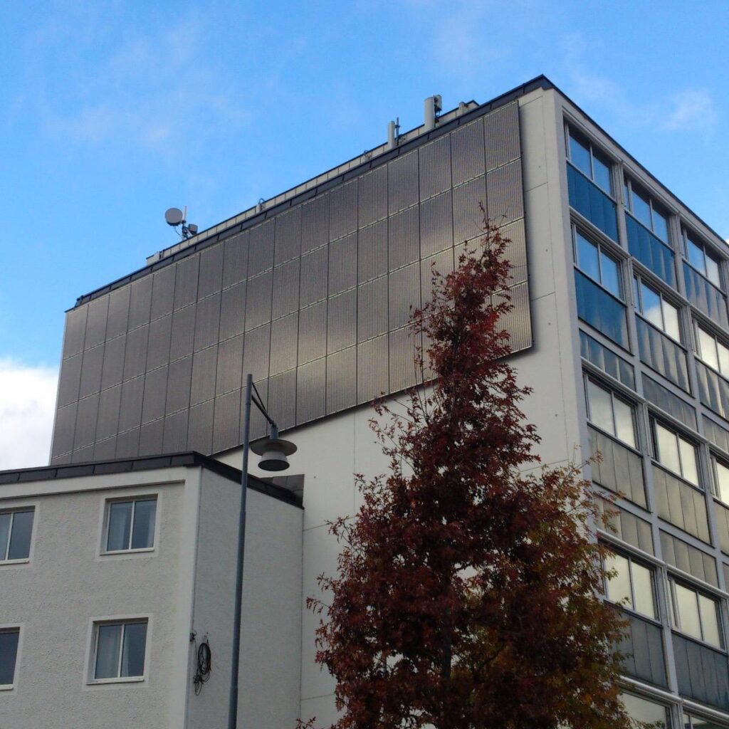 Hög byggnad med solpaneler på den övre fasaden. Ett träd med höstlöv och en gatlykta står i förgrunden, under en delvis molnig blå himmel.