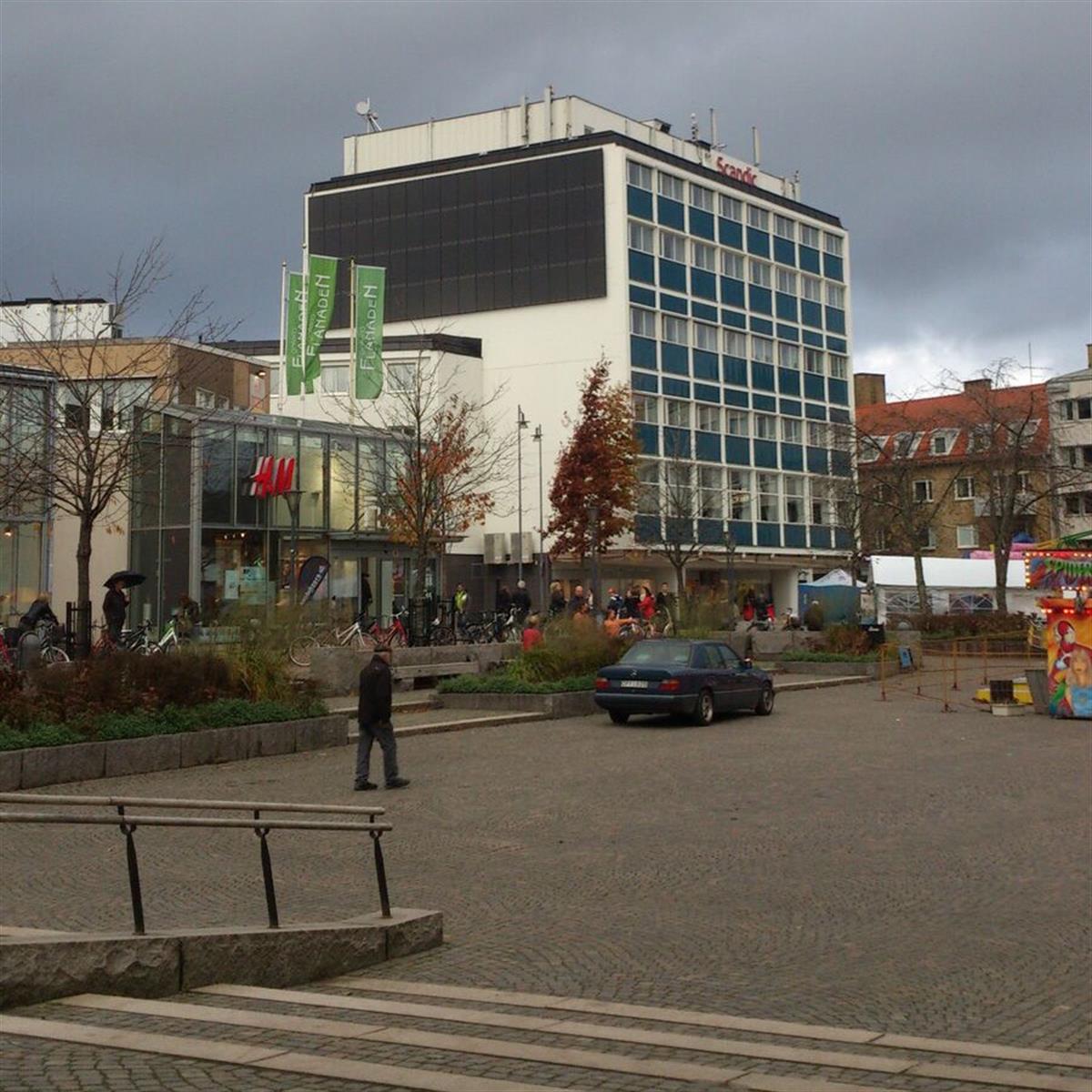 Ett stadstorg med en byggnad i flera våningar med en stor glasfasad. Flera personer går och en bil står parkerad på det öppna området. Mulen himmel och lövlösa träd är synliga.