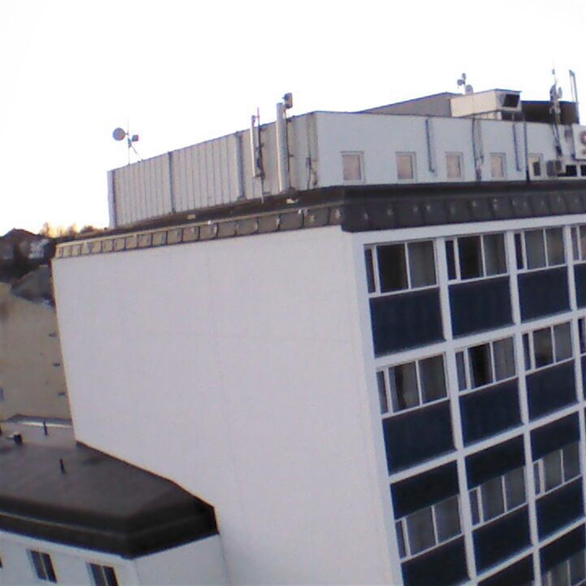 Vinklad bild av en vit flervåningsbyggnad med platt tak och blå panelsektioner. Takantenner och utrustning är synliga. Intilliggande strukturer och en klar himmel är i bakgrunden.