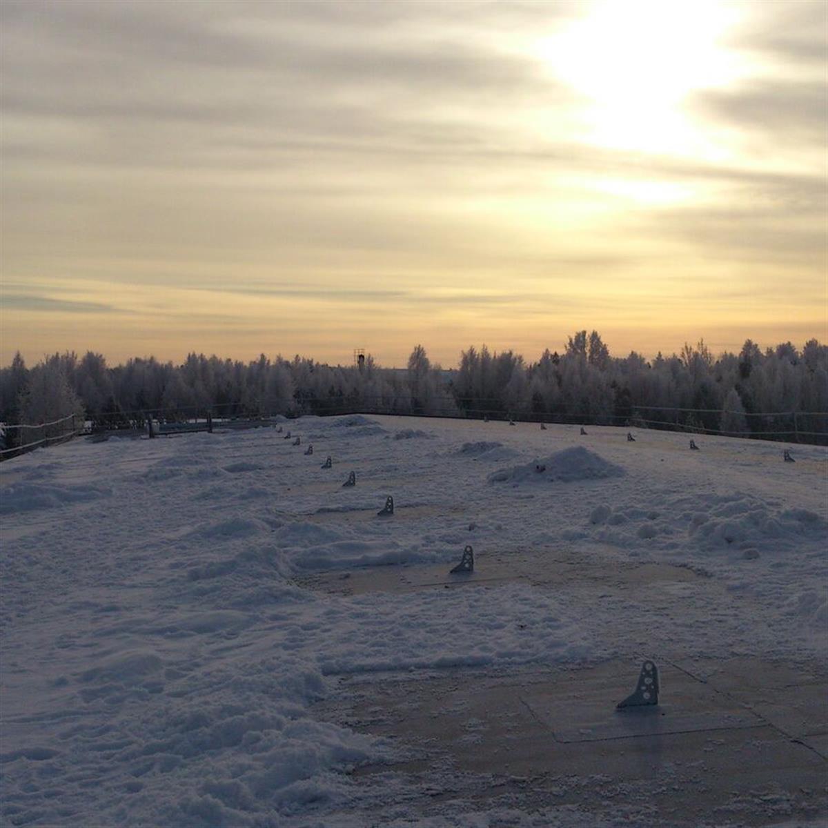 Snötäckt tak med snöskydd, med utsikt över en vintrig skog under en molnig himmel vid soluppgång eller solnedgång.