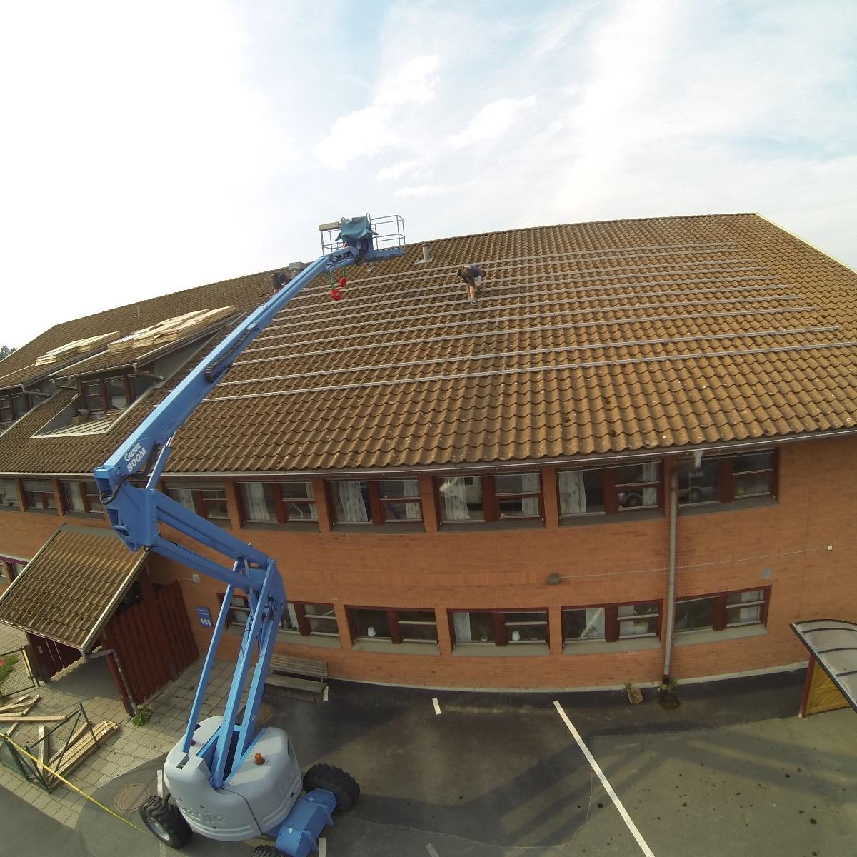 En person installerar solpaneler på ett stort tegeltak med hjälp av en körsbärsplockare, med en klar himmel i bakgrunden.