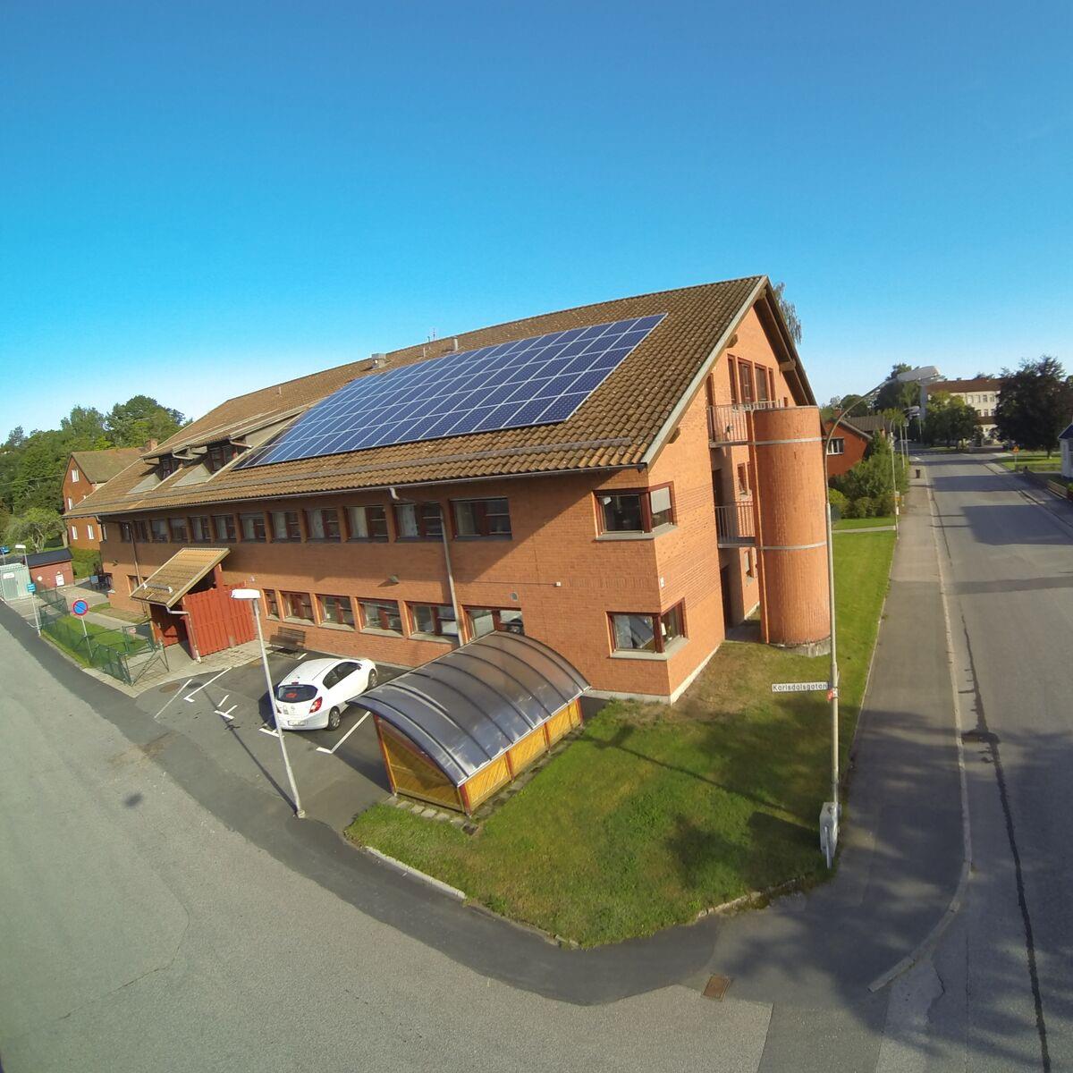 Flygfoto över en rött tegelbyggnad med solpaneler på taket, i anslutning till en väg och en parkeringsplats med en parkerad vit bil.