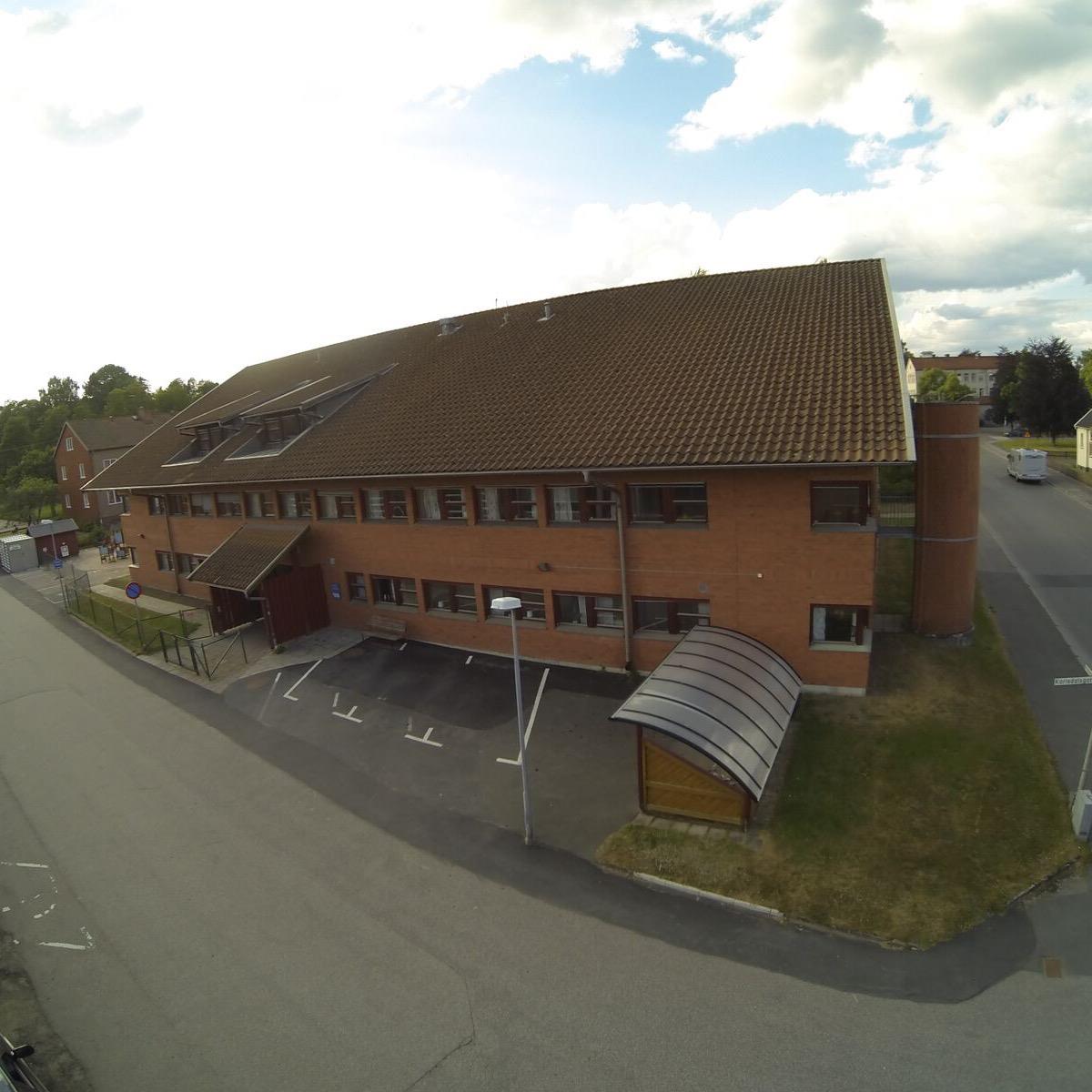 Flygfoto över en tvåvånings tegelbyggnad med tegeltak och flera fönster, bredvid en gata med några parkerade bilar.