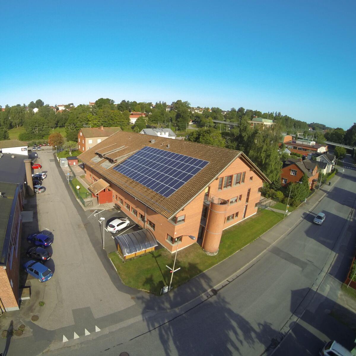 Flygfoto över en tegelbyggnad med solpaneler på taket, omgiven av träd och en parkeringsplats med bilar, belägen i en korsning med vägar och trottoarer.