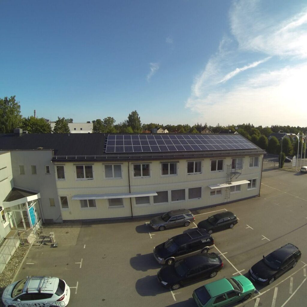 Flygfoto över en byggnad med solpaneler på taket, omgiven av parkerade bilar på en parkeringsplats, under en klarblå himmel.