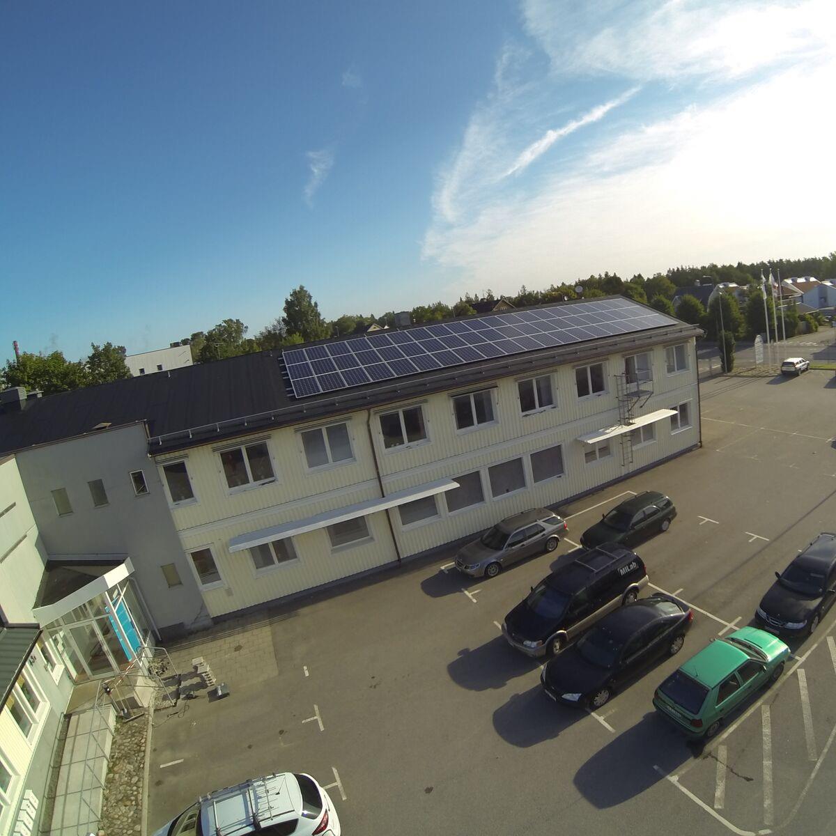 Byggnad med solpaneler på taket, omgiven av en parkeringsplats med bilar. Himlen är klar med få moln.