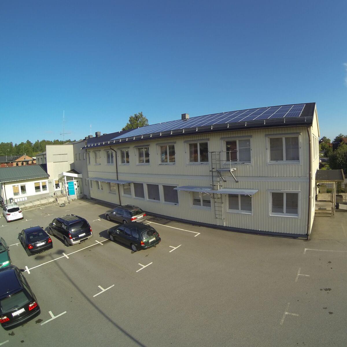 Tvåvånings beige byggnad med solpaneler på taket, omgiven av en parkeringsplats med flera parkerade bilar under en klarblå himmel.
