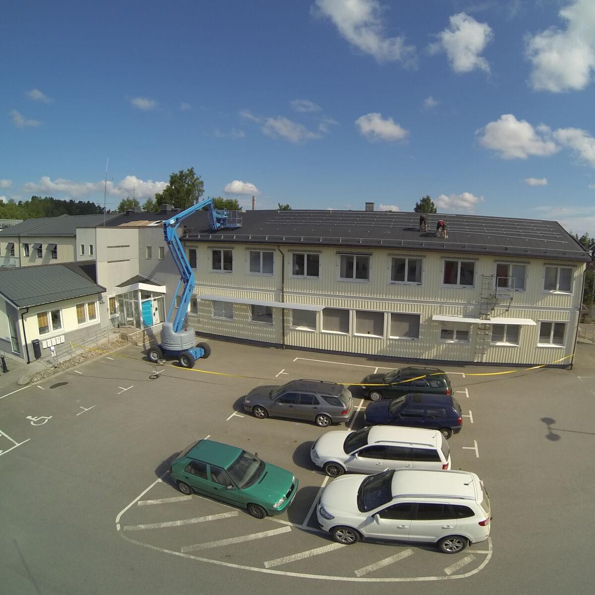 En blå hiss bredvid en tvåvåningsbyggnad med en arbetare på taket som installerar utrustning. Flera bilar står parkerade på intilliggande tomt.