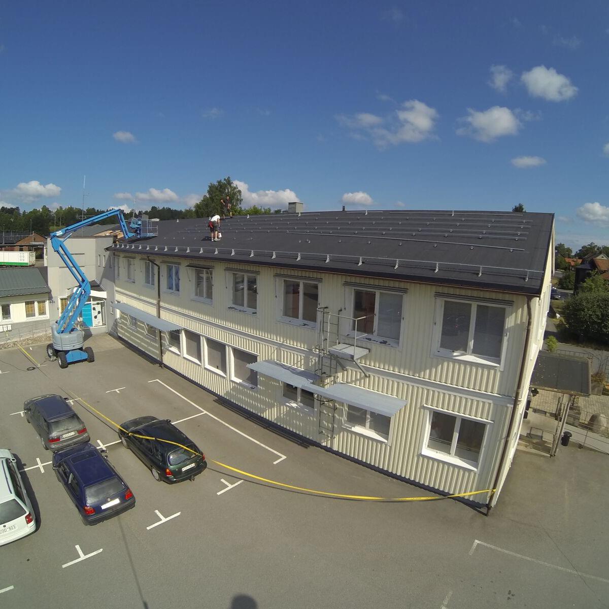 En person arbetar på ett tak och installerar solpaneler på en tvåvåningsbyggnad, med en hydraulisk hiss i närheten. Bilar står parkerade på tomten nedanför.