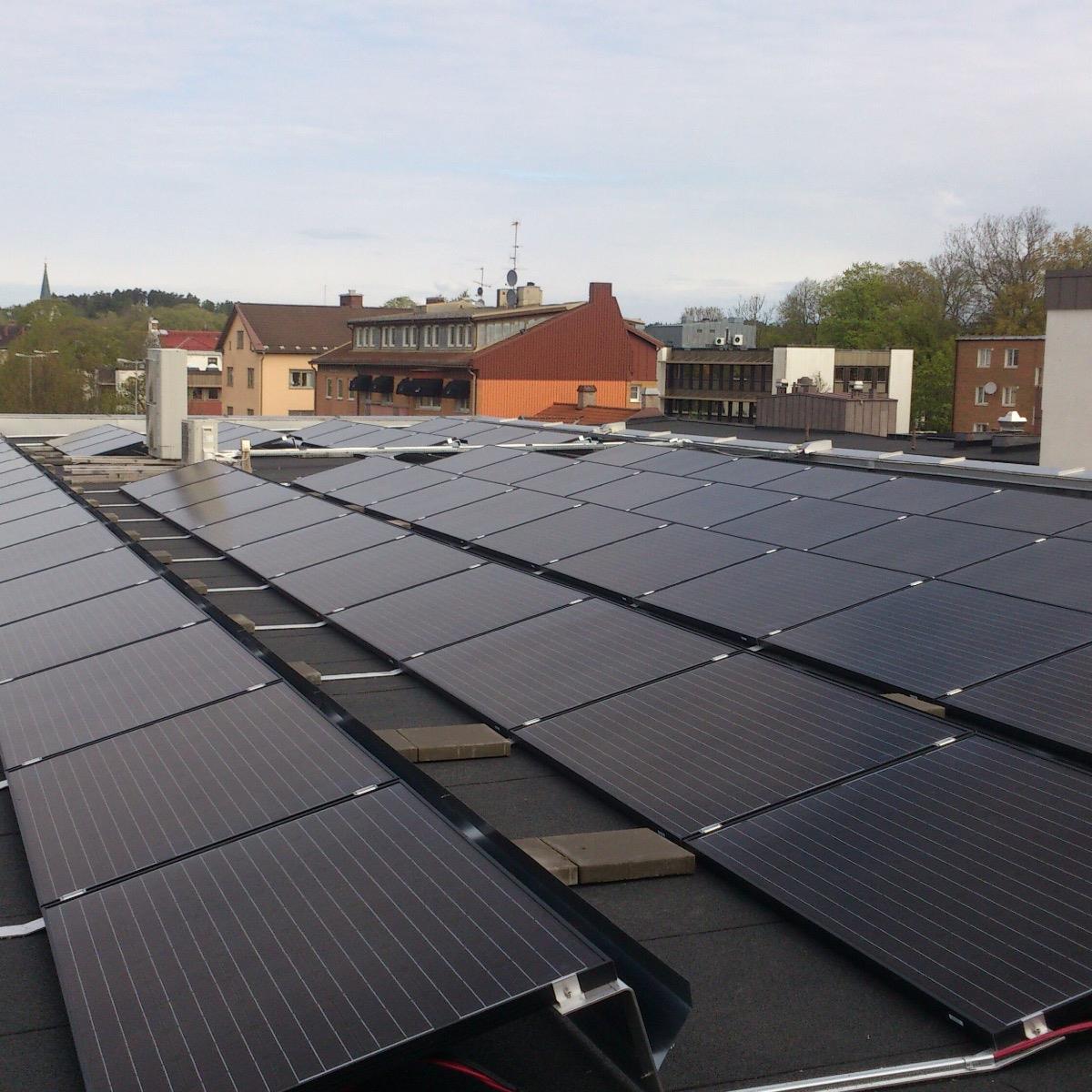 Solpaneler installerade på ett platt tak, med ett förortsområde och träd synliga i bakgrunden under en molnig himmel.