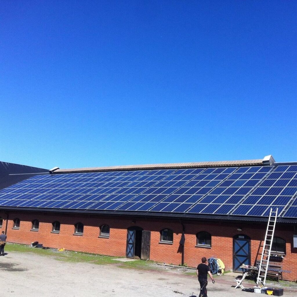 Tegelbyggnad med många solpaneler installerade på taket; två personer som står på marken i närheten mot en klarblå himmel.