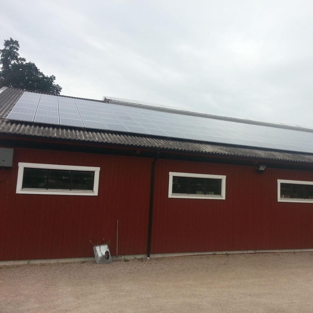 Röd byggnad med korrugerat tak med solpaneler. Mulen himmel och en skottkärra nära byggnaden.
