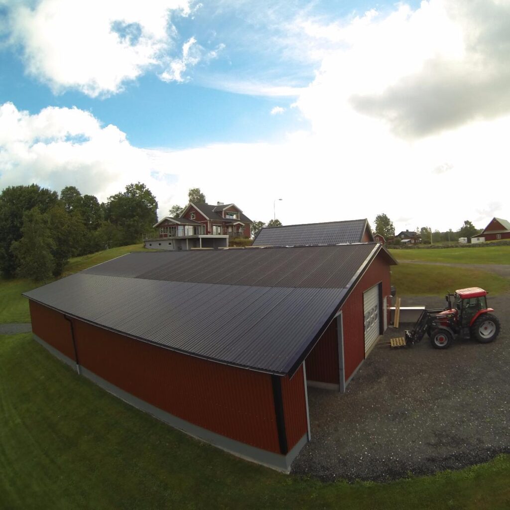 Röd lada med solpaneler på taket, belägen nära ett hus och träd, med en traktor parkerad utanför.