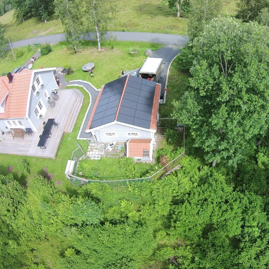 Flygfoto över två hus med solpaneler på ett tak, omgivna av träd och grönska. En väg kurvor i bakgrunden.