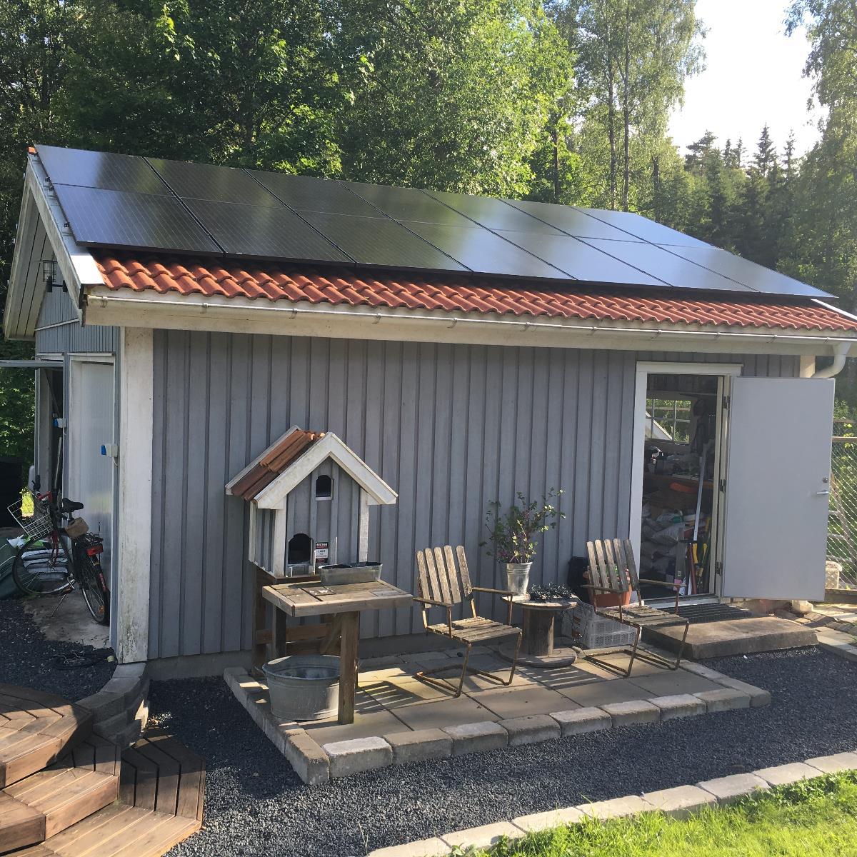 Liten grå bod med solpaneler på taket, öppen dörr som avslöjar lagrade föremål inuti. Däck med stolar, bord och en fågelholk. Omgiven av grönska.