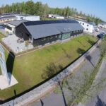 Flygfoto över en modern svart byggnad märkt Four Office omgiven av en grön gräsmatta, träd och en stenmur, med en parkeringsplats i närheten på en klar dag.