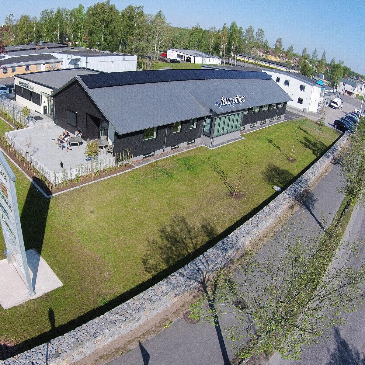 Flygfoto över en modern svart byggnad märkt Four Office omgiven av en grön gräsmatta, träd och en stenmur, med en parkeringsplats i närheten på en klar dag.