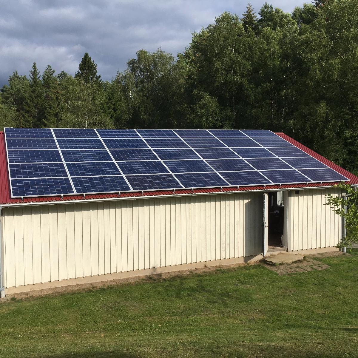En byggnad med rött tak har en rad solpaneler, omgiven av grönt gräs och träd under en molnig himmel.