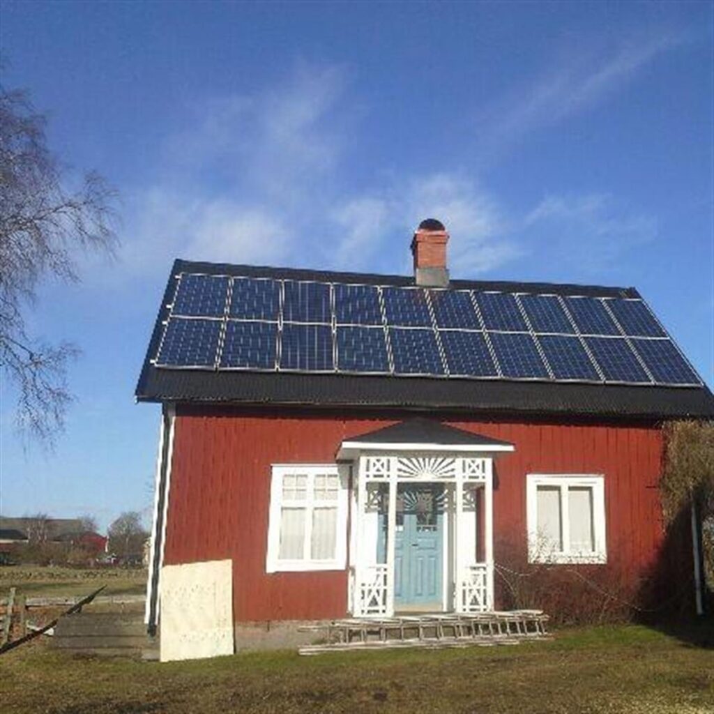 Ett rött hus med en blå dörr och vit klädsel har solpaneler installerade på taket. En skorsten är synlig och himlen är klar.