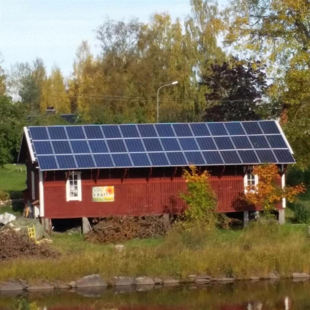 Röd träbyggnad med solpaneler på taket, omgiven av träd och gräs, med en liten damm i förgrunden.