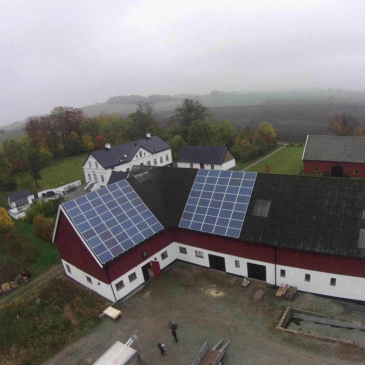 Flygfoto över en lantgård med solpaneler installerade på taken av två stora lador. Runt gården finns åkrar och träd under en molnig himmel.