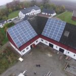 Flygfoto över en lantlig fastighet med solpaneler installerade på taken av två sammankopplade byggnader, omgiven av åkrar och träd.