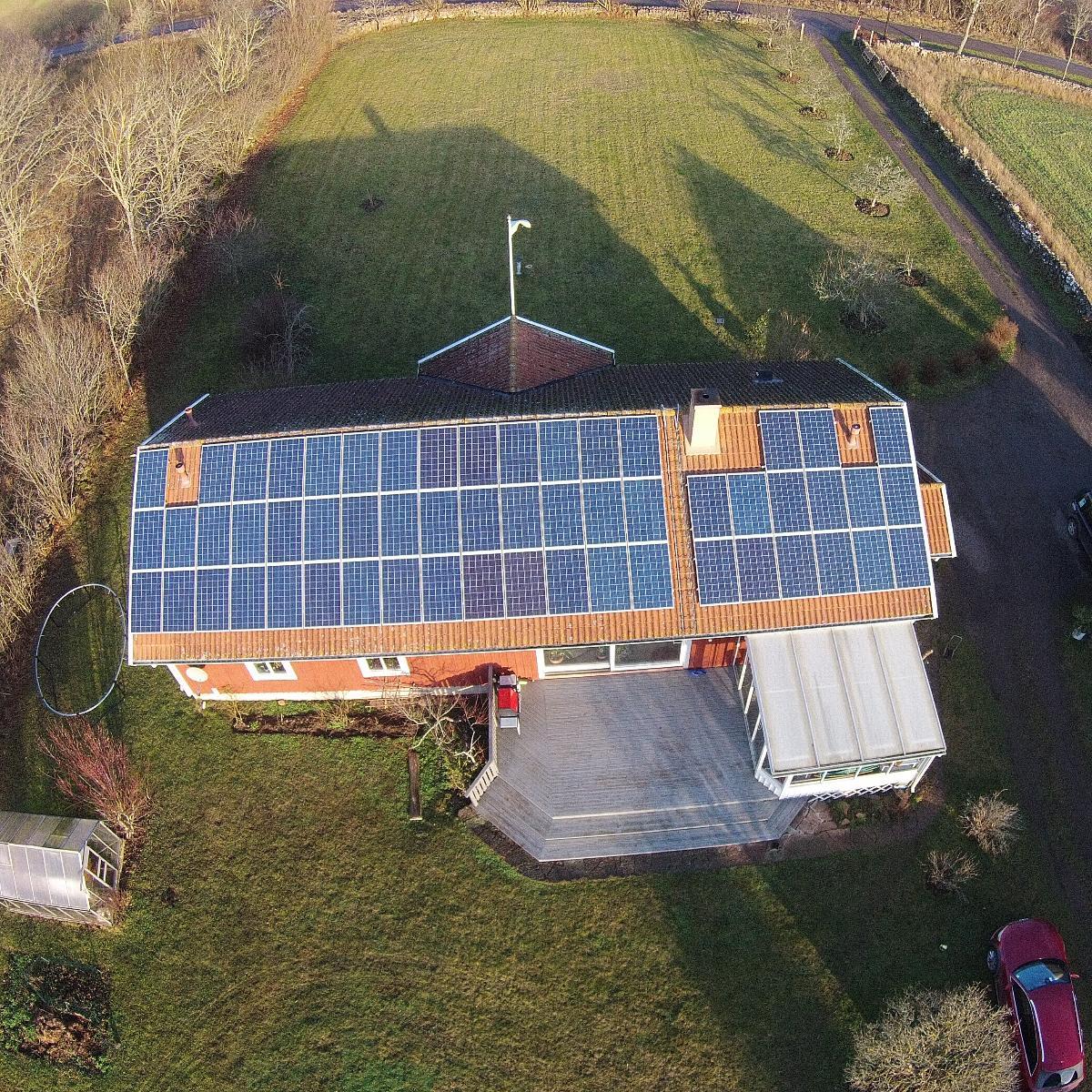 Flygfoto över ett hus med solpaneler som täcker taket. Till fastigheten hör en uteplats och ett litet växthus, omgivet av gräs och några träd.