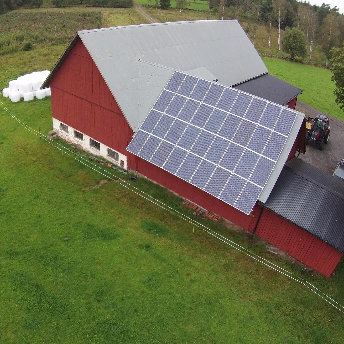 En röd ladugård med solpaneler på taket, belägen i ett gräsområde med träd i bakgrunden.