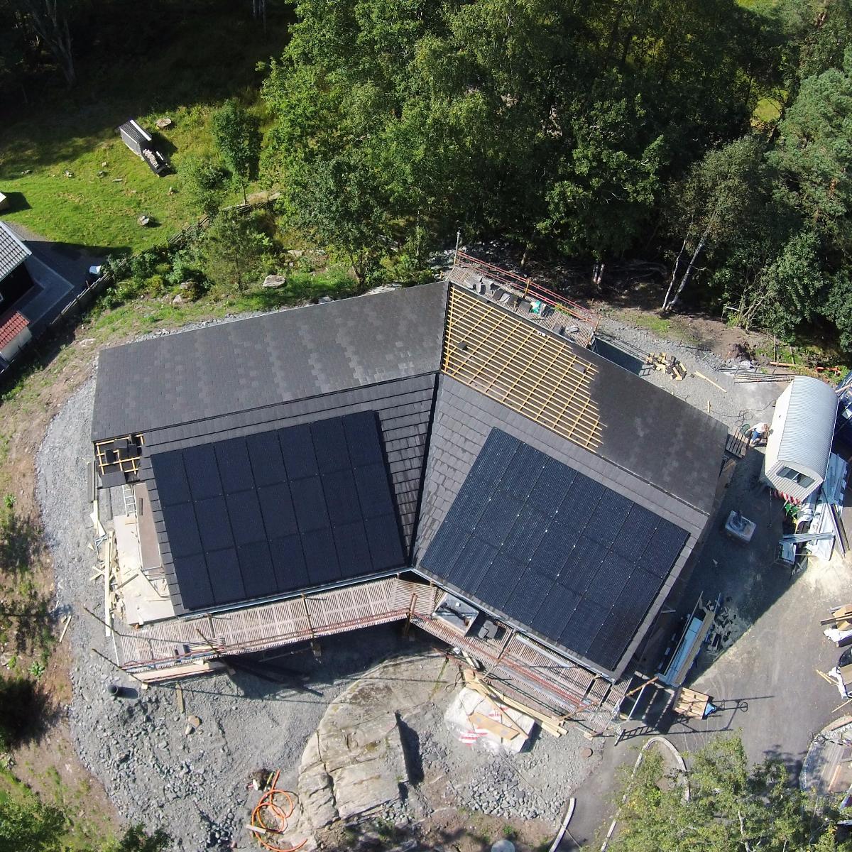 Flygfoto över ett hus under uppbyggnad med solpaneler installerade på taket. Omgivningen är täckt med grus och byggmaterial. Träden syns i bakgrunden.